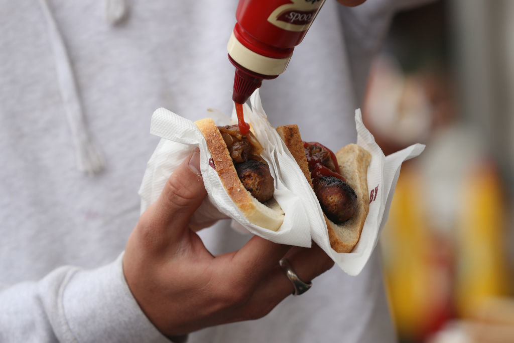 Sausages laid onto two slices of white bread with fried onions on top, and a ketchup bottle squeezing sauce onto them.