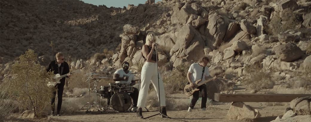 A full band plays in the desert in front of a rock formation with Hanna behind the microphone