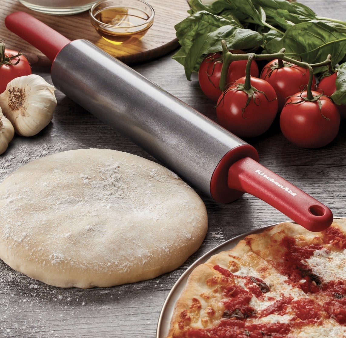 The silver and red rolling pin is surrounded by a pizza and the supplies to cook another