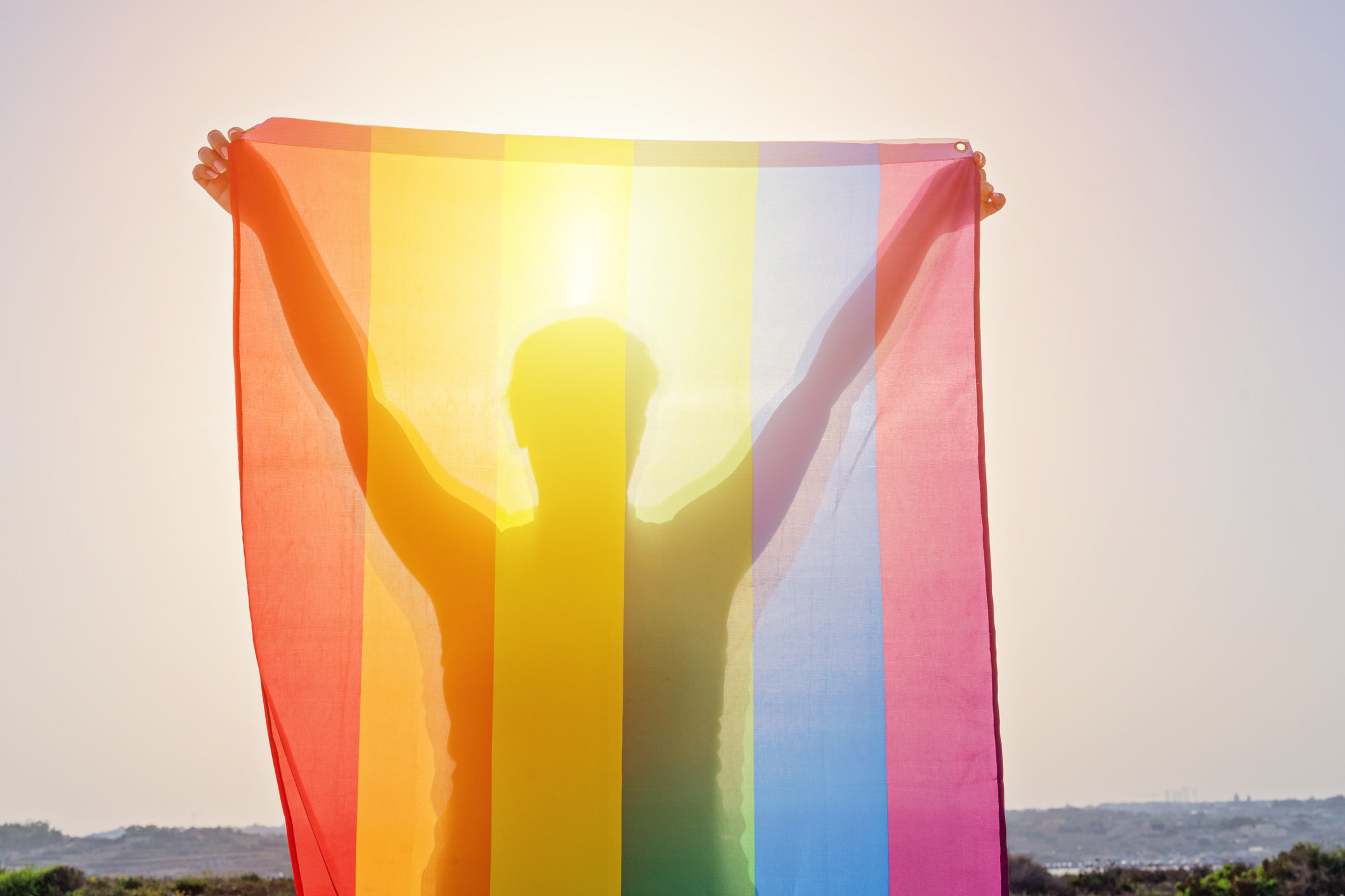 Young woman holding raised hands waving LGBT rainbow flag against sky