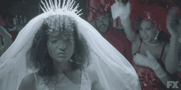 Angel in a wedding veil with spiky crystalline headpiece, big, curly hair, and in wedding gown, posing with her hand under her chin in front of sparkly partiers. 