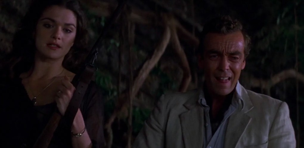 Evie and Jonathan stand next to each other in a jungle; Evie holds a gun