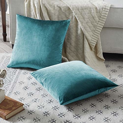 Two teal cushion covers on the floor beside a sofa with a cream throw blanket and some books 