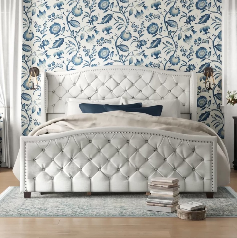 An antique white, tufted bed with navy throw pillows in front of a navy, paisley wallpapered wall 