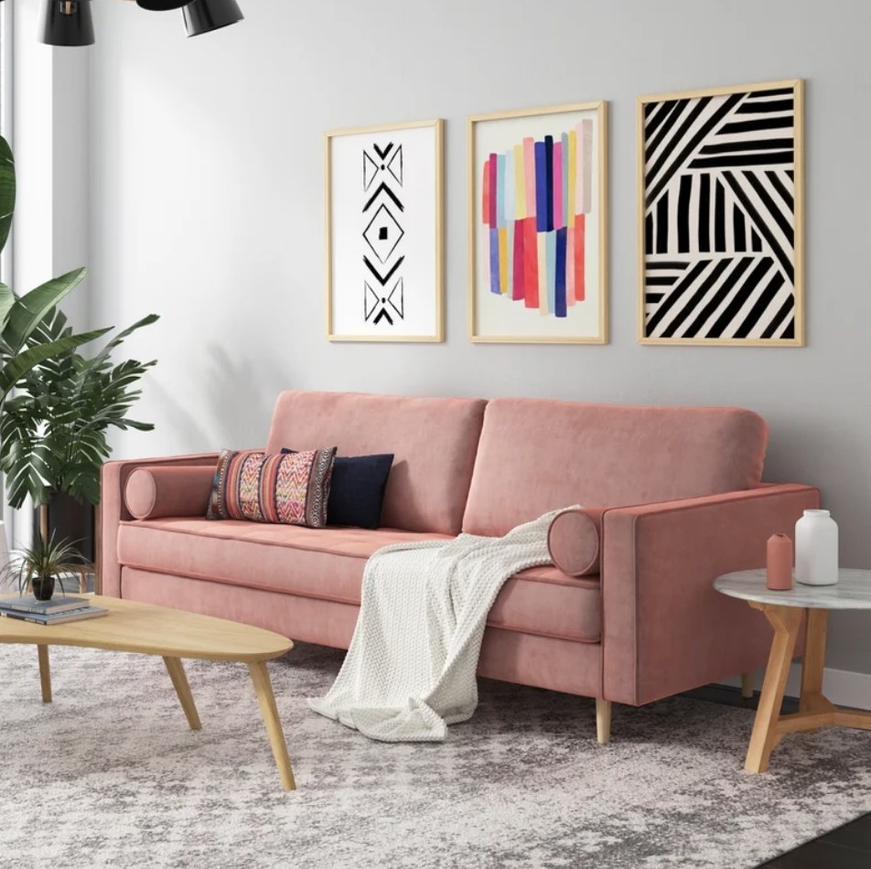 A blush pink, velvet sofa displayed in a living room with accent pillows and a throw blanket on top