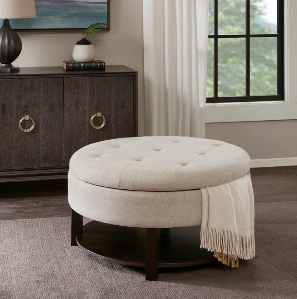 A cream, round, tufted storage ottoman with a blanket inside displayed in a living room