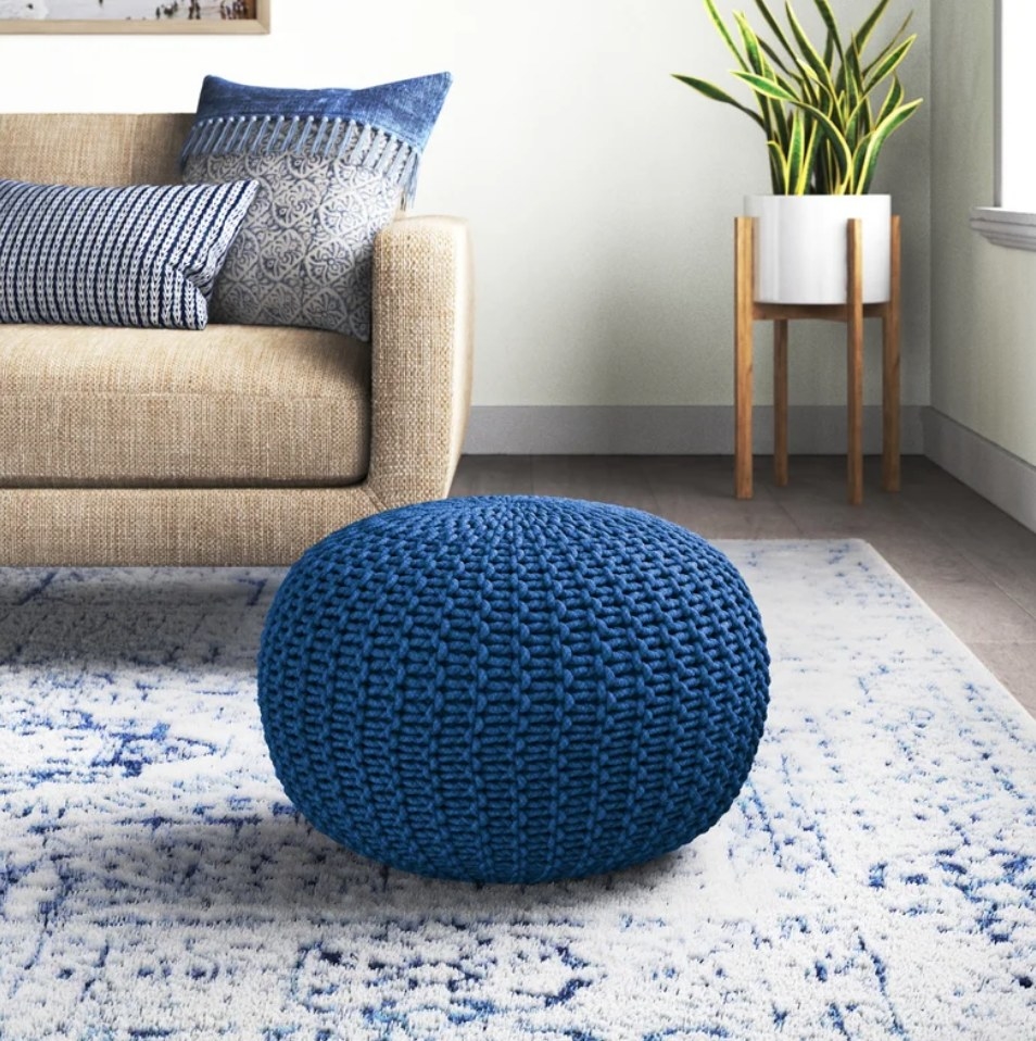 A cotton, round cobalt pouf on a carpet in front of a couch with matching blue accent pillows