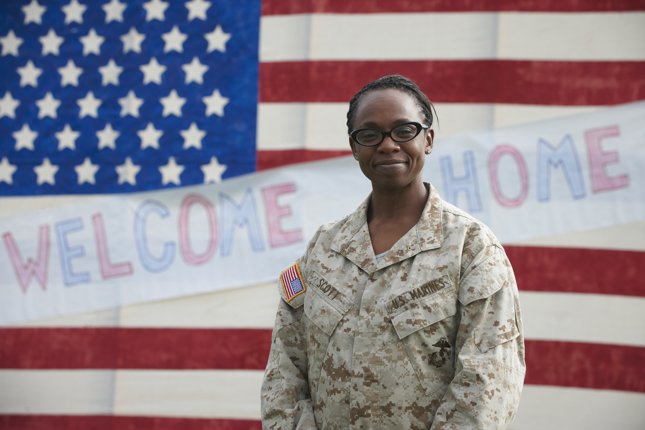 Female veteran standing in front of an American flag with a paper sign on it that says, &quot;Welcome home&quot;