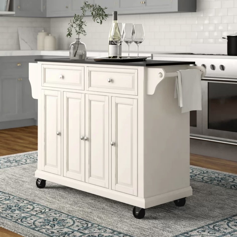 A white kitchen island on wheels with 2 racks, 2 drawers 4 doors, and 3 cabinets with a solid black granite countertop 