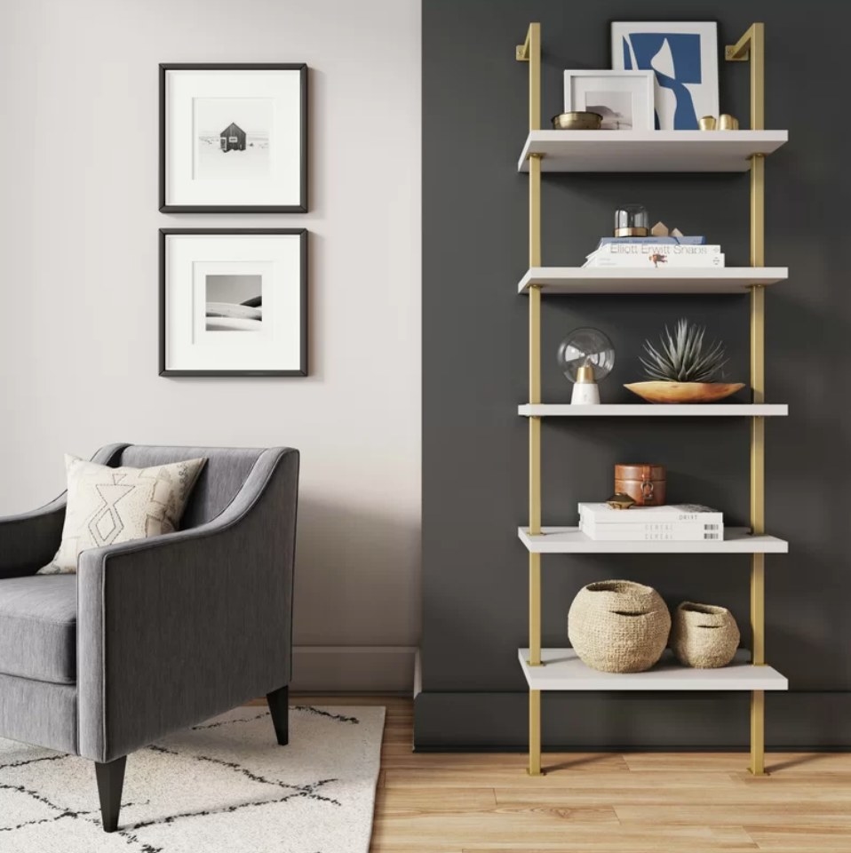 A gold/white steel ladder bookcase filled with chic decor pieces next to an accent chair on a carpet
