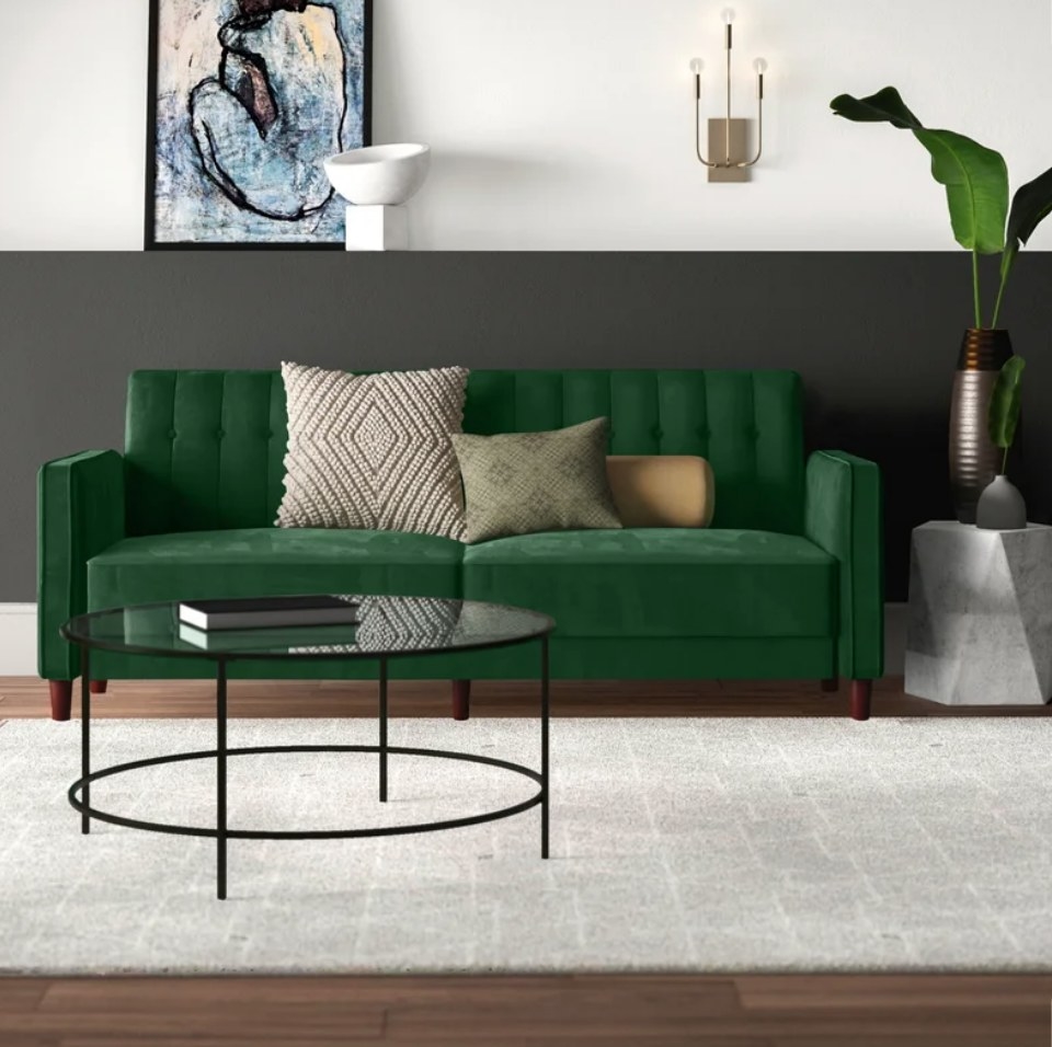 A green velvet sleeper sofa with throw pillows displayed in a living room behind a glass and steel coffee table