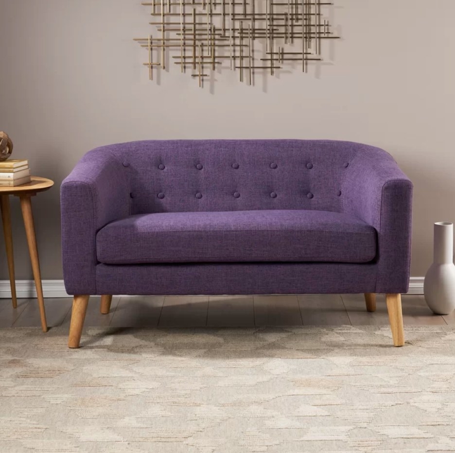 A purple, polyester loveseat with wooden legs displayed under modern wall art