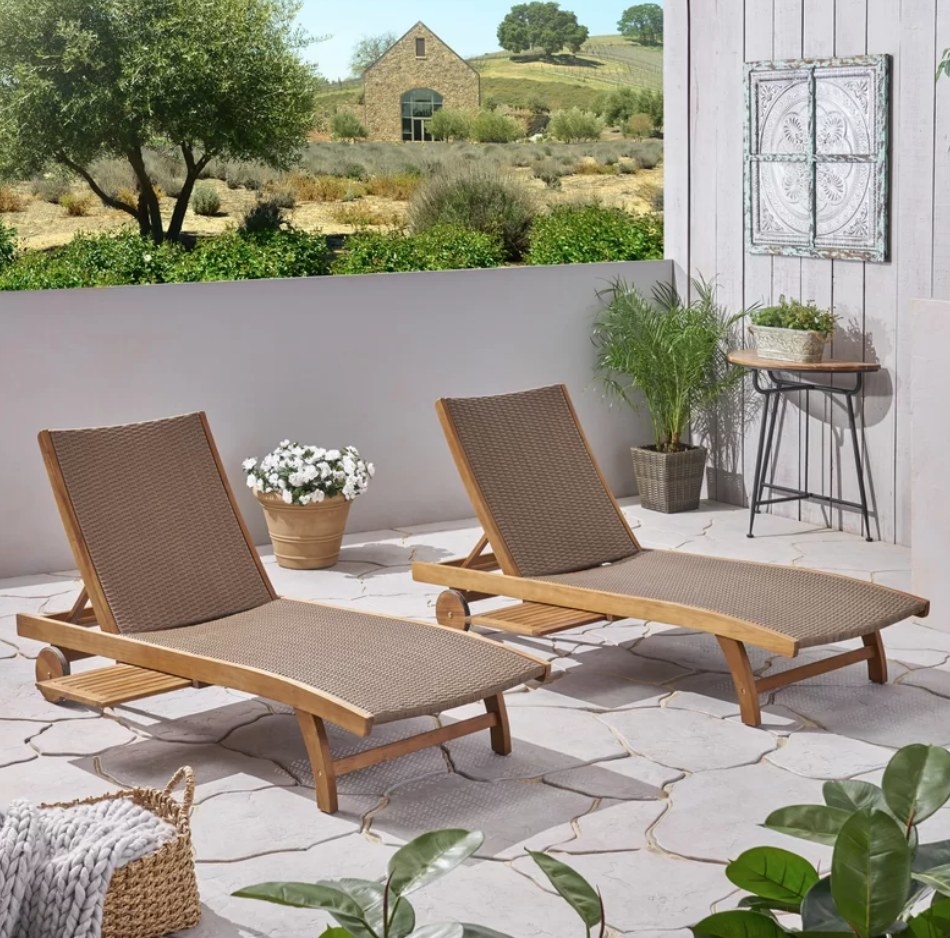 A set of two teak and brown outdoor, reclining chaise lounge chairs on a patio with built in pull-out side trays