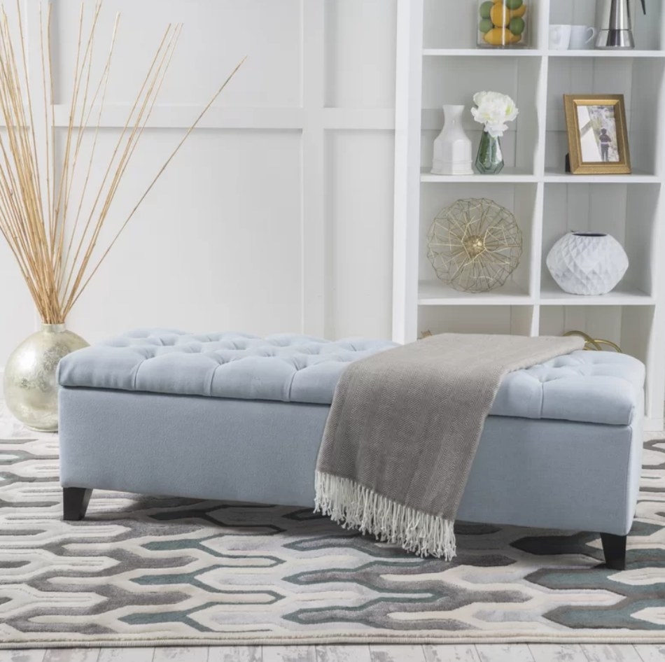 A light blue tufted storage bench with a grey throw blanket draped over