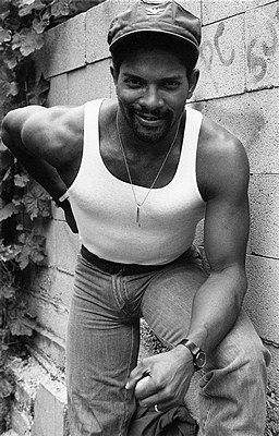 A Black man in jeans and a tank top and a hat leans forward to the camera
