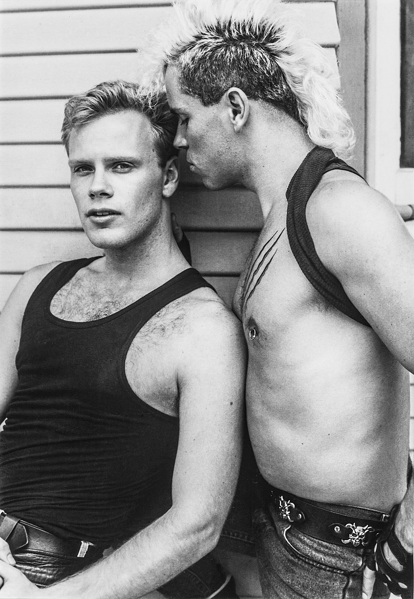 A shirtless man with a mohawk hairdo whispers in the ear of another man
