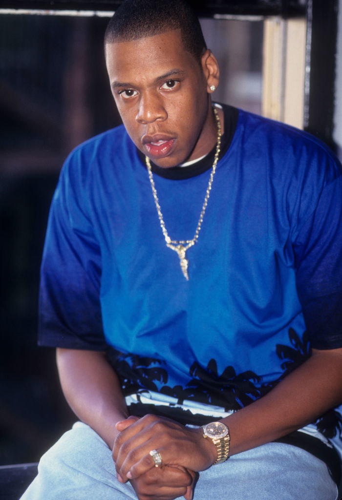 Jay-Z crossing his hands and wearing a blue shirt and a chain as his photograph is taken