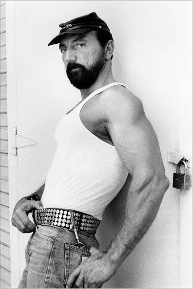 A bearded white man in jeans and a tank top leans against a door