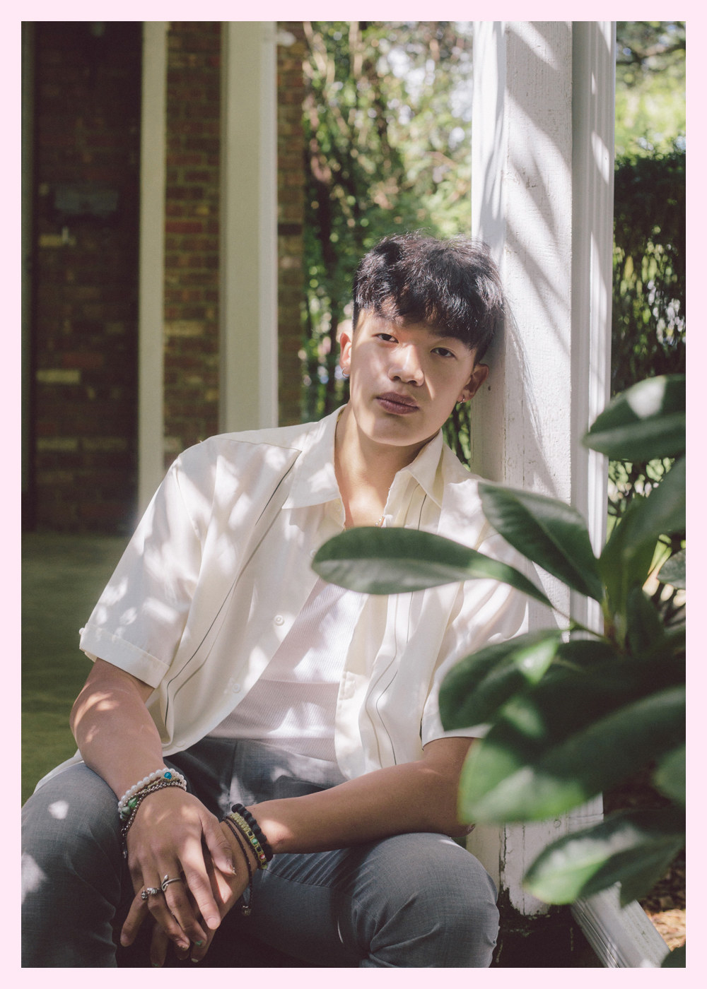 A young man in a white shirt with jewelry leaning against a post outside a home, with a plant in the foreground 