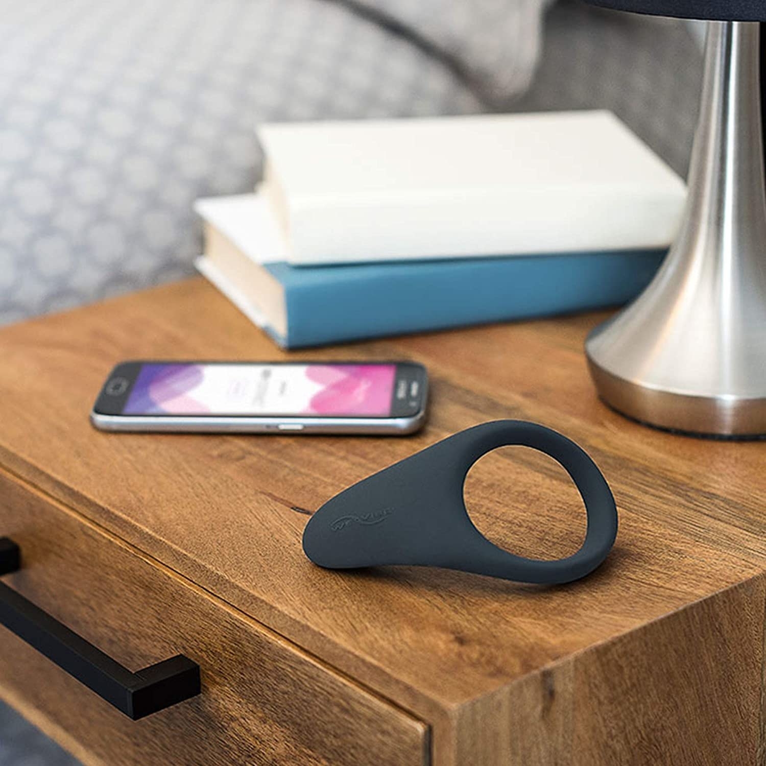 the cock ring on a bedside table with books and a smartphone displaying the app