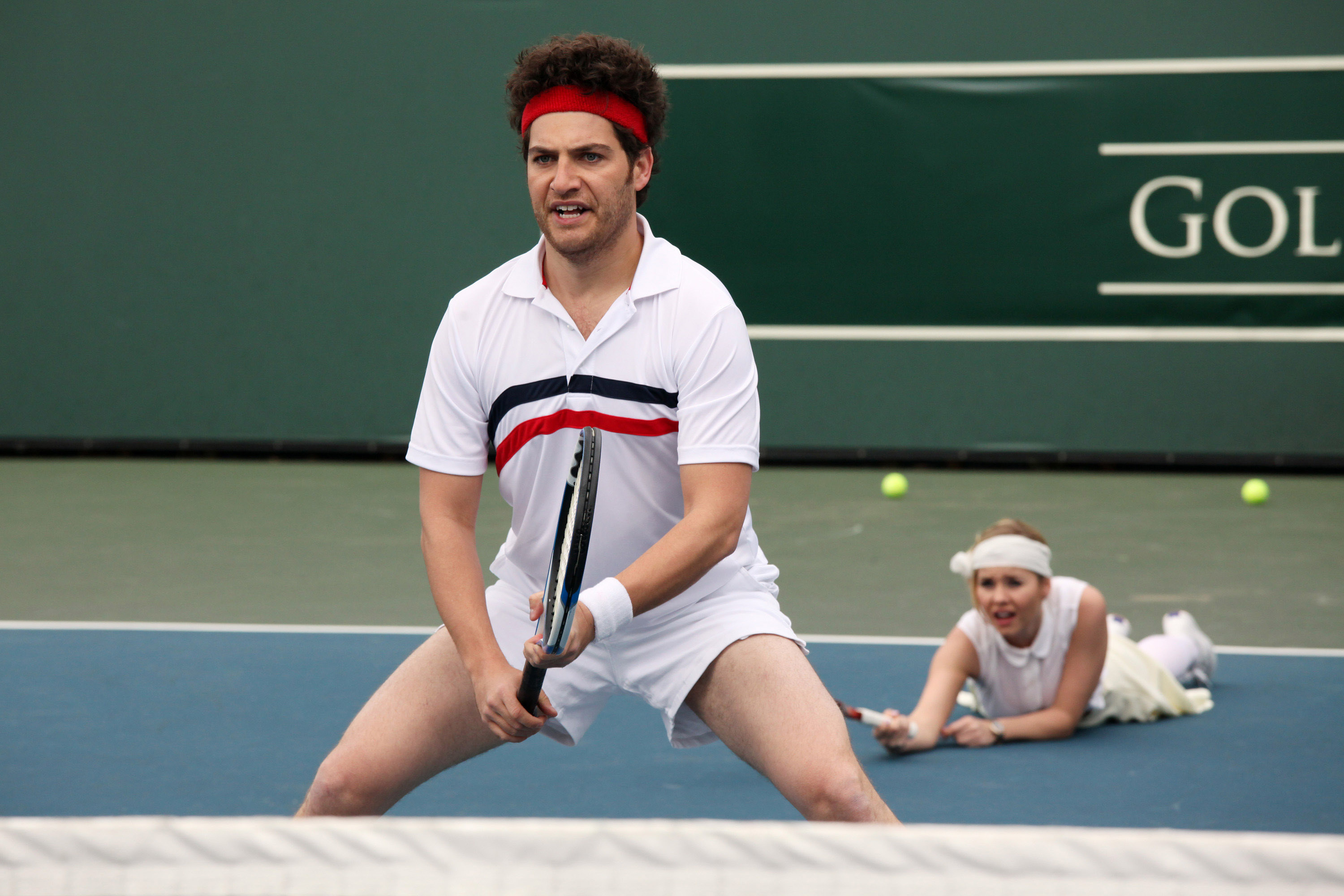 Adam Pally plays tennis with Elisha Cuthbert on the ground behind him