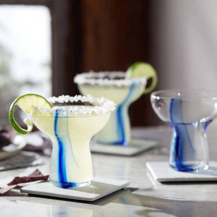 three stemless margarita glasses with blue ribbons blown into the glass. Glasses are filled with margaritas with limes and salt on the rim.