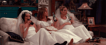 Monica, Rachel, and Phoebe in Friends sit on a couch together wearing wedding dresses and drinking