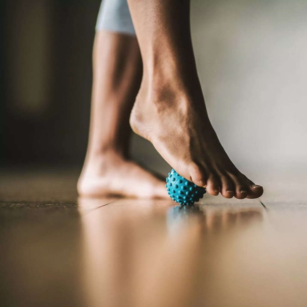 A model using a Gaiam blue body massager under their foot against the floor