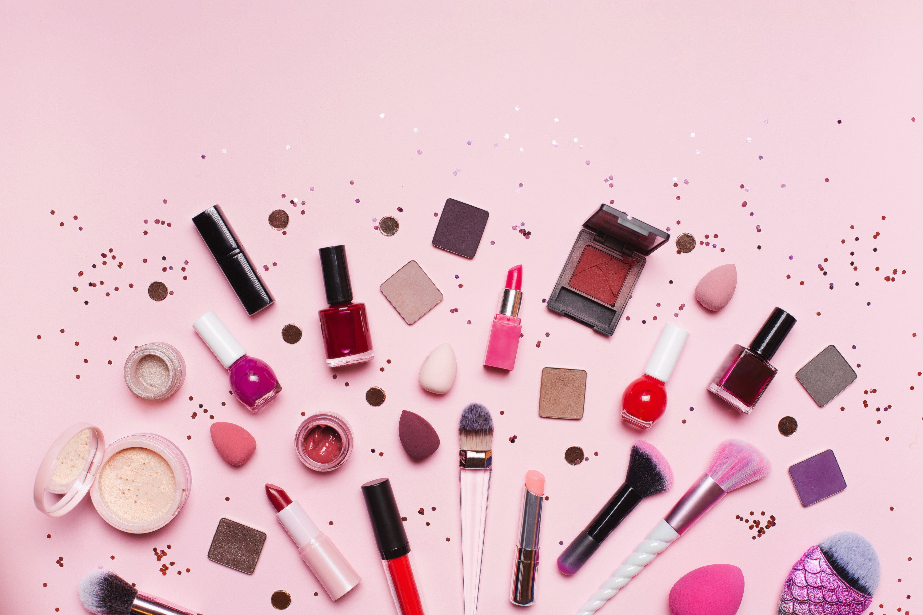 set of assorted tools and products for makeup application placed near shiny glitter on pink background