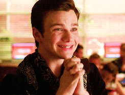 Kurt Hummel smiles brightly as he claps his hands together on his chest and jumps up and down.