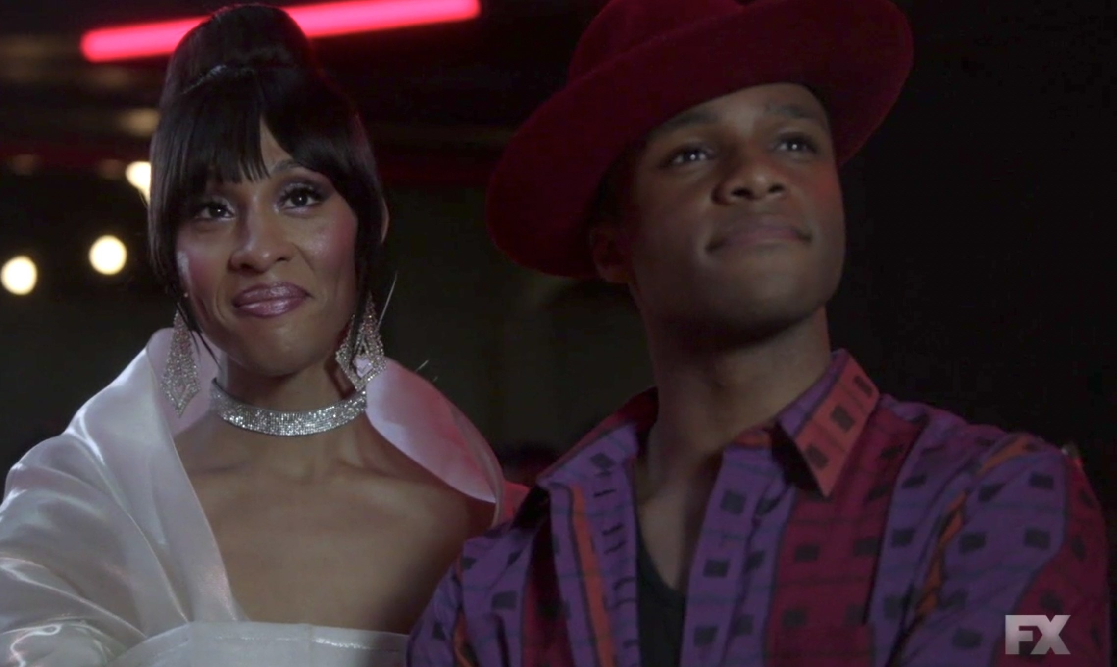 Blanca in a delicate shawl and sparkling collar necklace and earrings with an updo, next to Ricky in a wide brim hat and patterned button down shirt with lines and squares