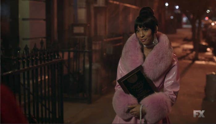 Blanca in a coat with wide fur cuffs and collar and an updo, holding her legendary mother award standing on the sidewalk looking towards a group of people. 