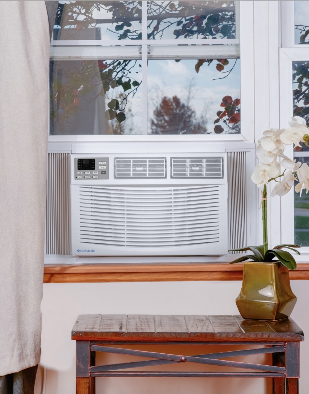 The white window air conditioner is in an open window and surrounded by a tree and flowers