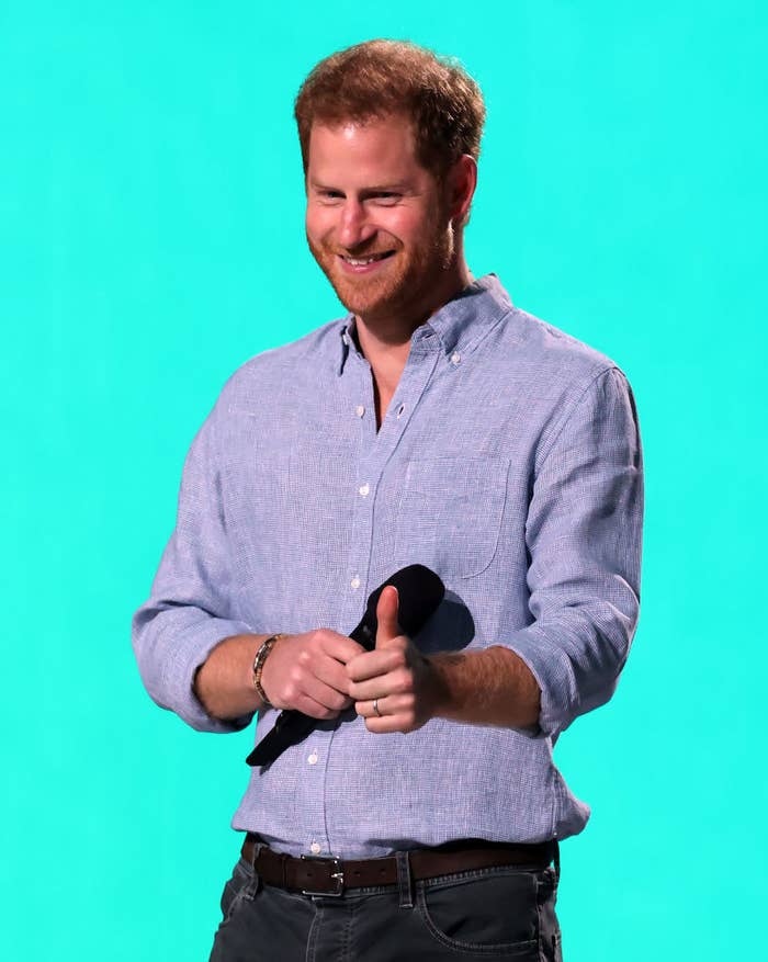 Prince Harry smiles on stage while giving a thumbs up, dressed in a blue shirt and holding a microphone