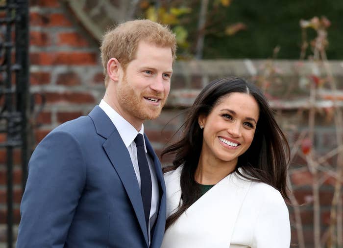 Prince Harry, wearing a blue suit, smiles for the camera while posing next to wife Meghan Markle, who is wearing a white coat