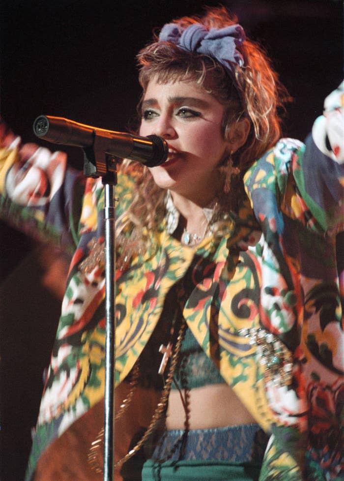 Madonna performing in concert in 1985 while wearing multiple chain necklaces, a colorful jacket, and big hair