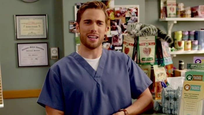 A chest up image of Ted Mullins wearing his blue vet scrubs and standing next to the front desk of his vet clinic. There are diplomas on the wall and pet accessories in the background.