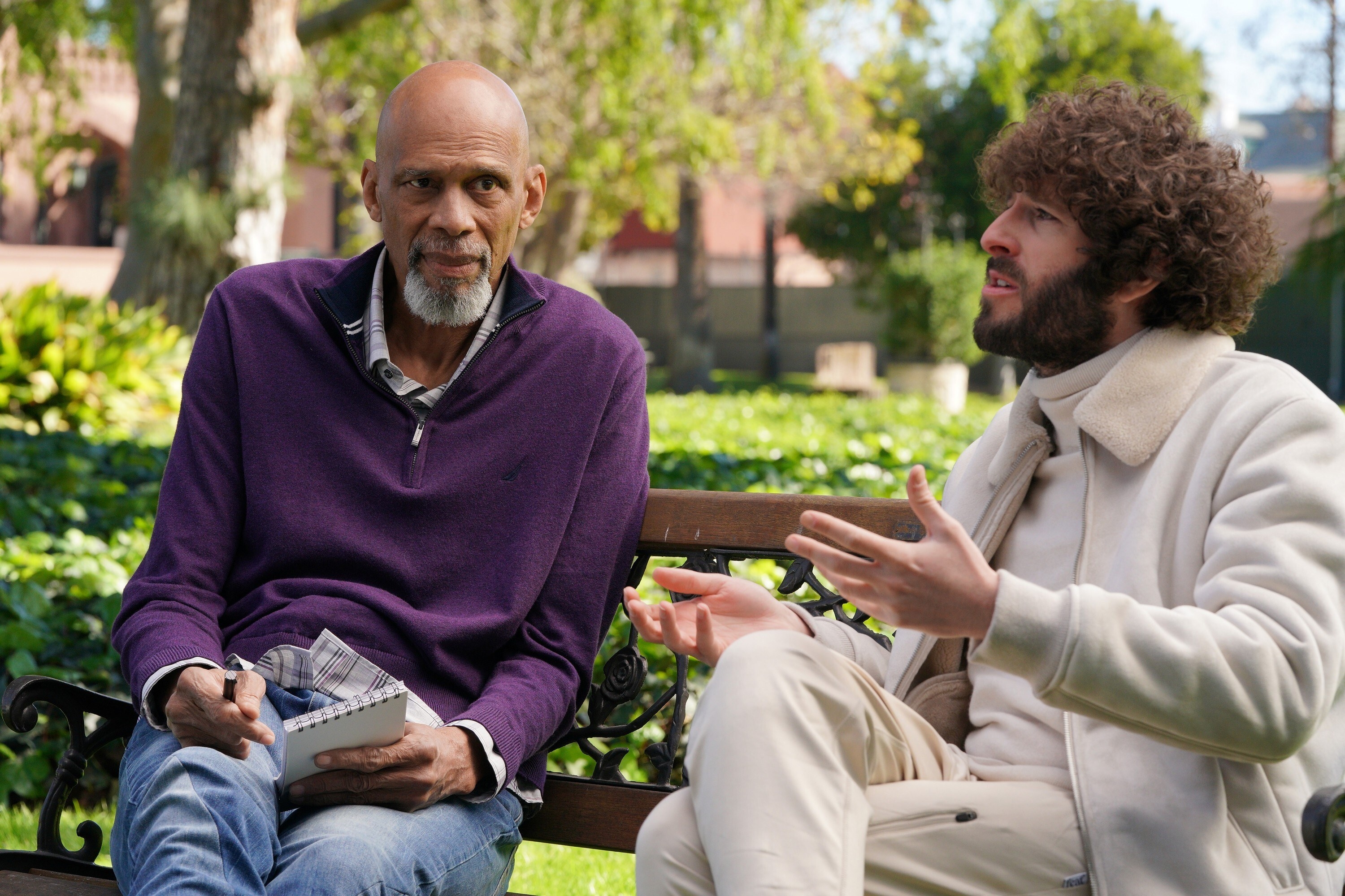 Kareem Abdul-Jabbar in sweater and jeans with notepad sitting with Dave in sweatsuit on a bench outside