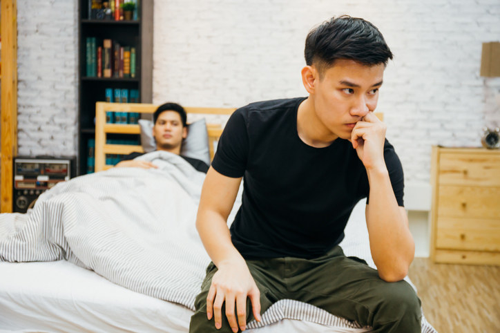Man sitting on the edge of the bed while his partner is lying in bed, both with frustrated expressions