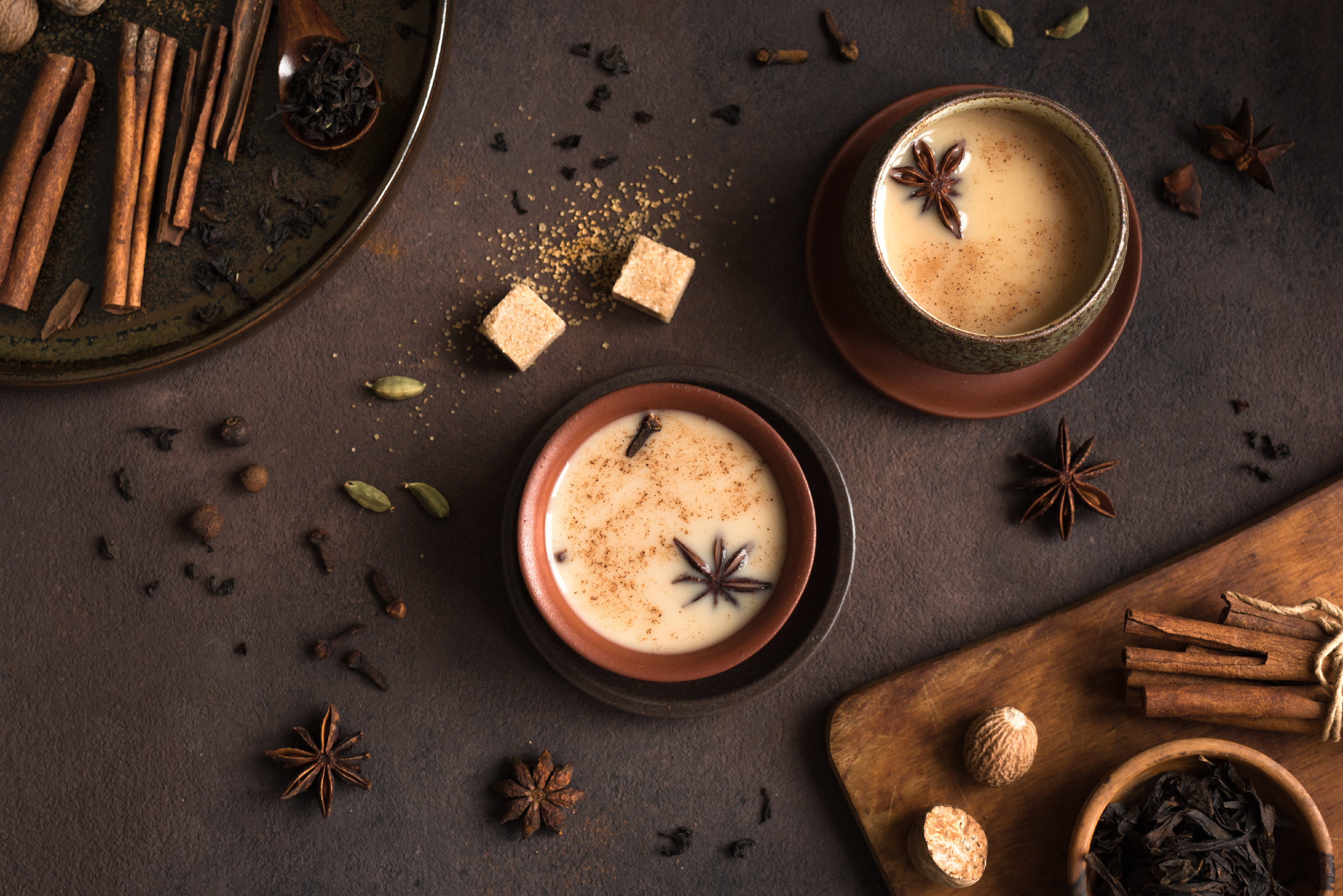 A cup of chai surrounded by ingredients like star anise and cinnamon sticks and contrasted against a dark grayish-purple background.