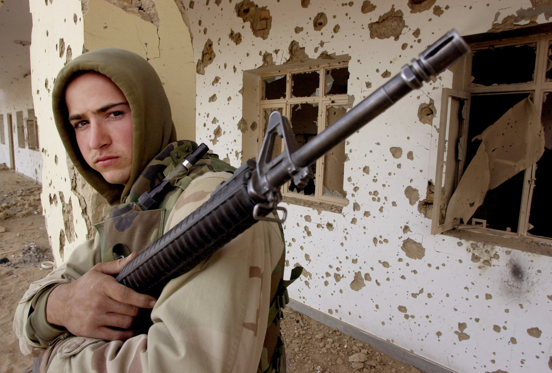 A young man stands with a gun in front of a bullet riddled building in Afghanistan