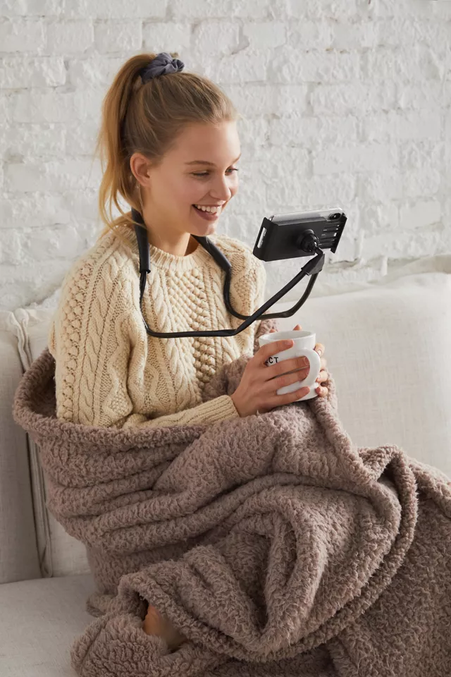 A person sitting on their couch holding a cup of coffee while watching something on their phone using the phone mount around their neck