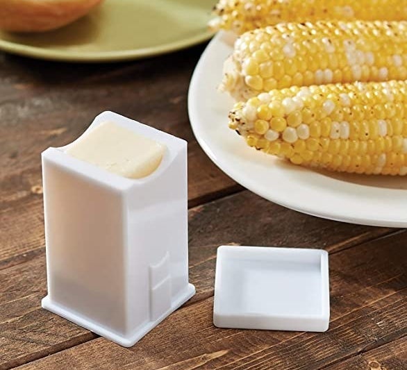 a white tube with a stick of butter inside next to a plate of corn