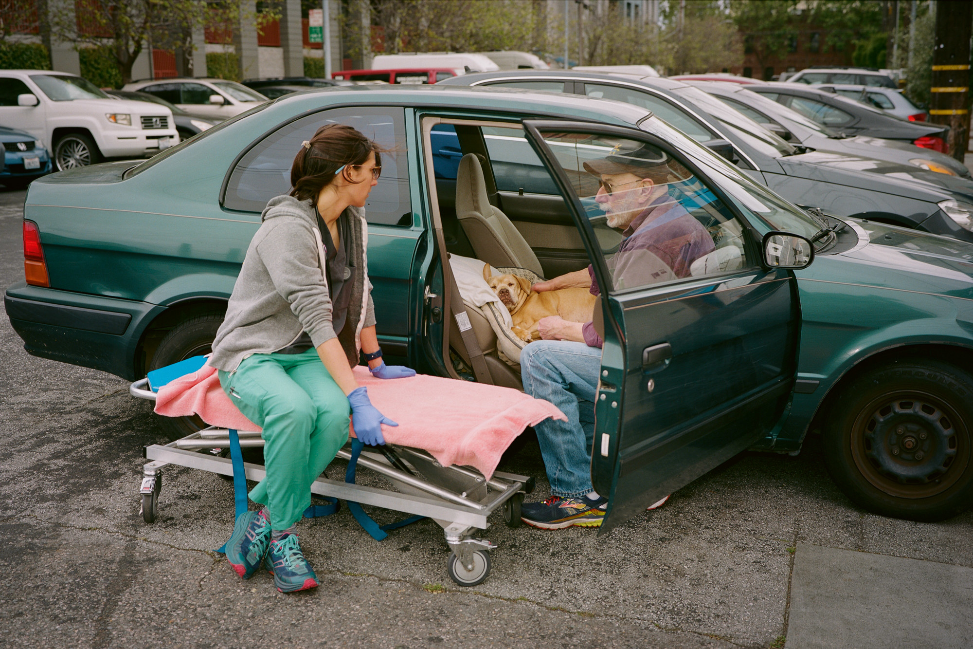 A veterinary nurse sits on a stretcher and talks to an older man who has his dog in the front seat of a car