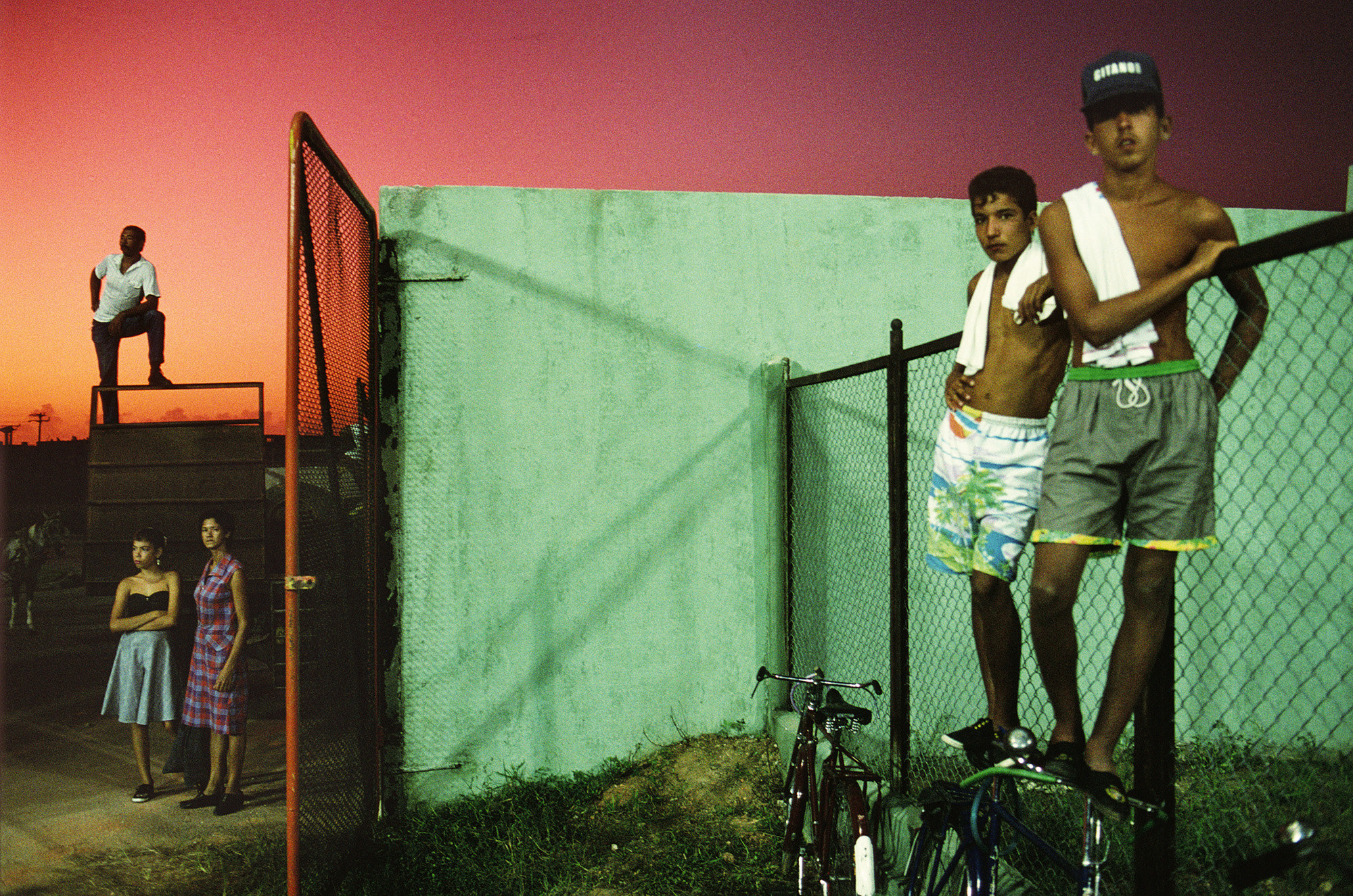 A man stands on a fence at left, two women stand below him, and on the right two young men stand on bikes by another fence