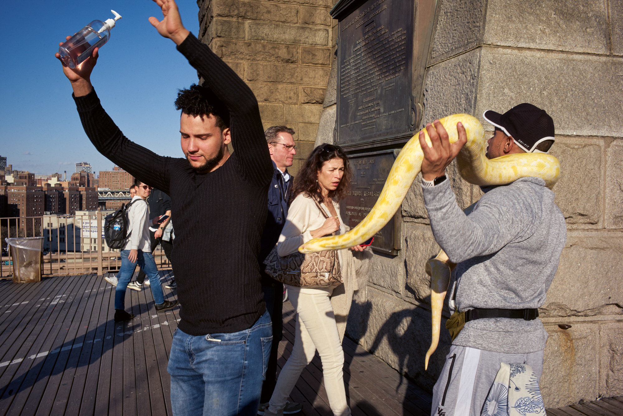 A man dances on the Brooklyn Bridge while a couple walks past, another man carrying a large snake