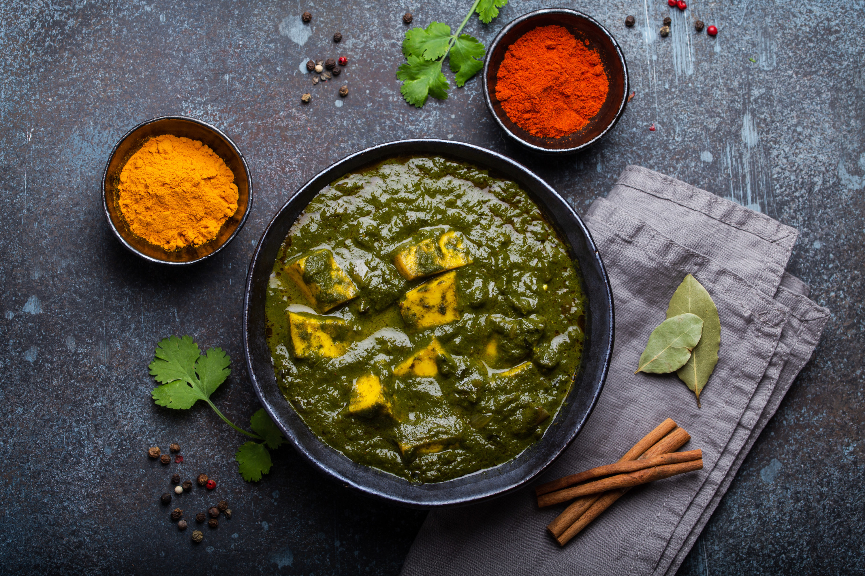 Palak paneer, which is green with white cottage cheese (paneer), is in a black bowl and on top of a light gray napkin. Spices, cilantro, cinnamon sticks, and herbs surround the bowl and are contrasted against a gray background