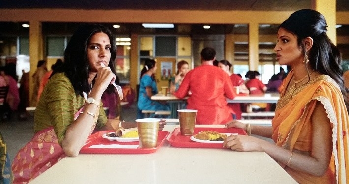 Devi&#x27;s mom and Kamala sit down at a cafeteria table to eat lunch. They each have a plate of food and a drink in a gold plastic cup a on a red tray