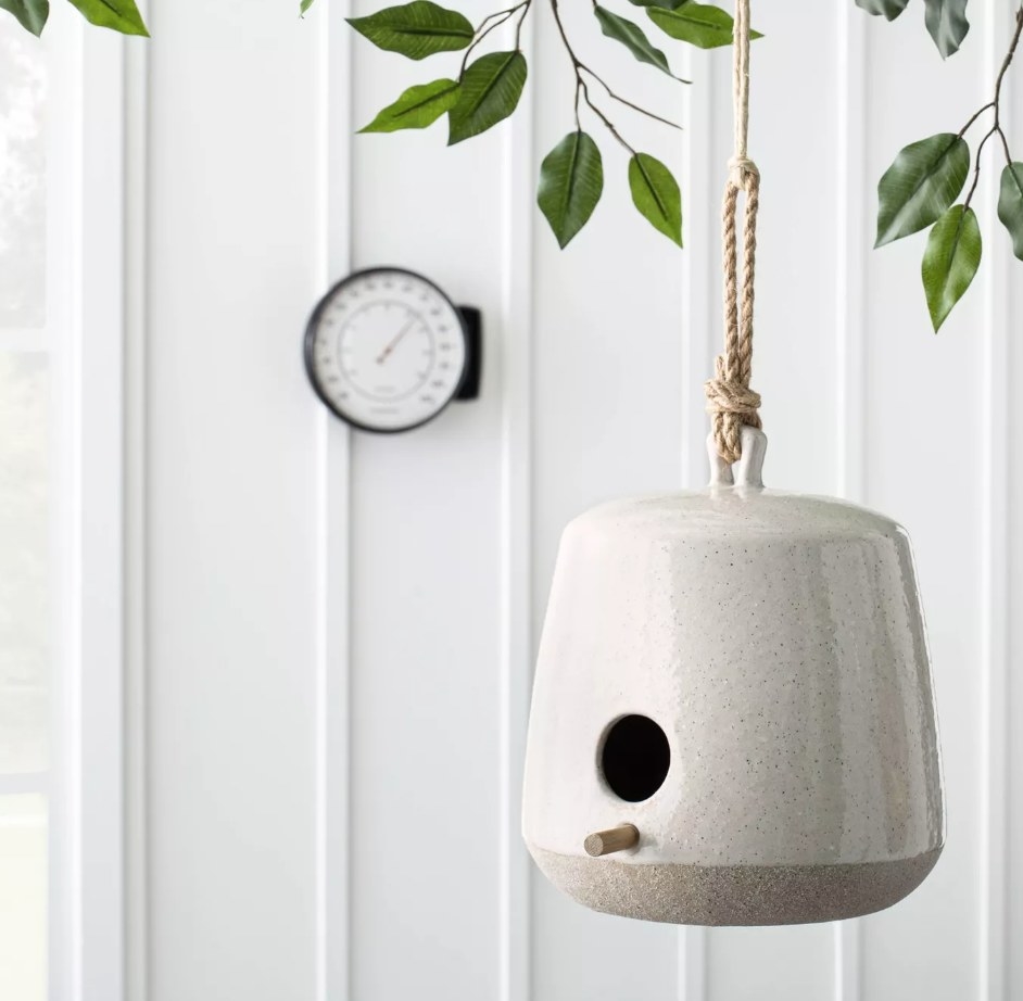 A speckled, stoneware bird house with a wooden perch hanging outdoors