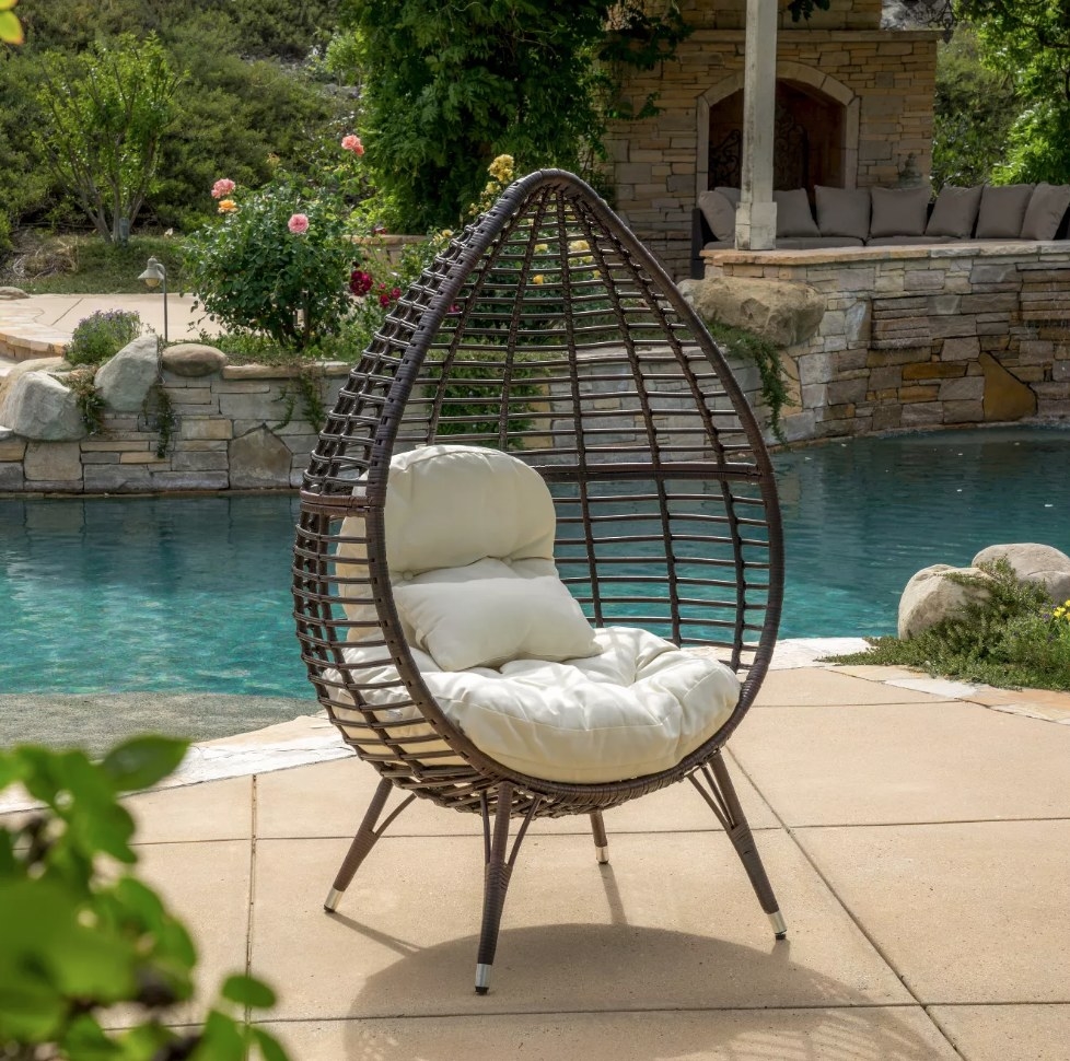 A brown wicker, teardrop egg chair with beige cushions on a deck near a pool